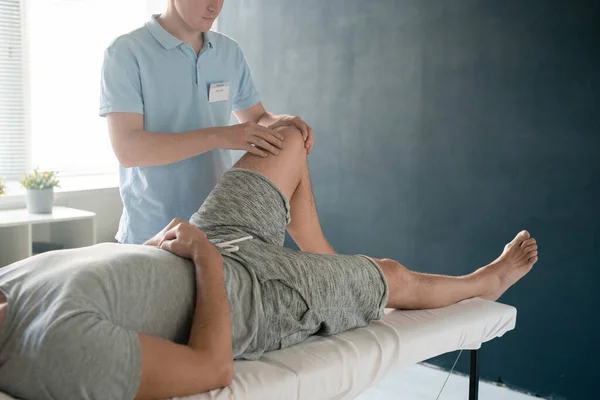 在康复过程中 年轻理疗师按摩弯曲左膝时 男性病人躺在医疗沙发上 — 图库照片
