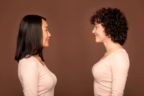 Side view of two cheerful young females of Asian and Caucasian ethnicities in white pullovers looking at each other over brown background