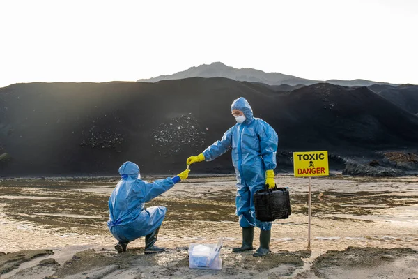 科学研究中に同僚の手から汚染水や土壌のサンプルを取り保護作業服の現代生態学者 — ストック写真