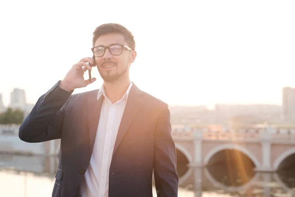 满足于现代的年轻胡子经理 戴着眼镜站在城市河边 用电话交谈 — 图库照片
