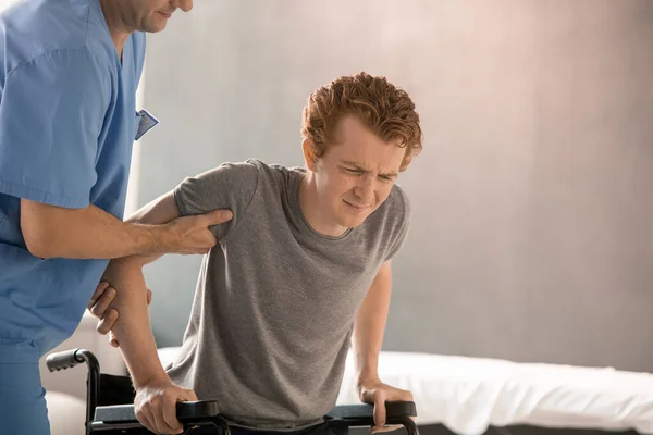 穿着制服的当代康复理疗师帮助年轻人在训练前从轮椅上下来 同时支撑他的胳膊和肘部 — 图库照片