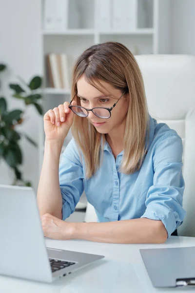 青いシャツを着たかなり自信のあるビジネスマンが 電子文書を扱う際に眼鏡の上にノートパソコンのディスプレイ上のデータを見ています — ストック写真