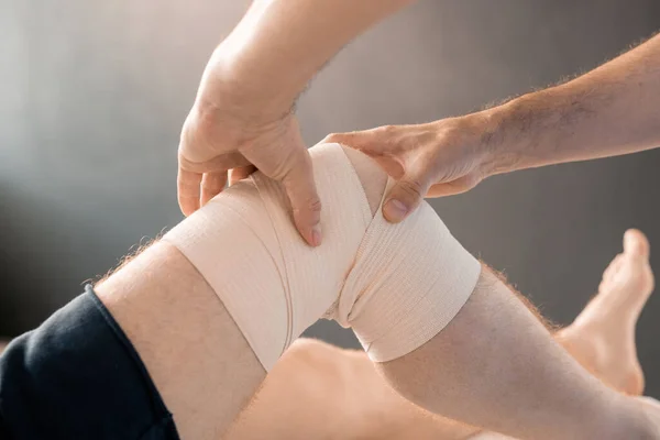 当代临床医生在康复过程中用软绷带包扎年轻男性患者膝部按摩的双手 — 图库照片