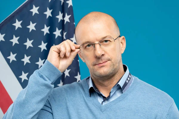 Orta yaşlı, kendinden emin bir avukat ya da işadamı kameranın önünde bayrağa karşı dururken gözlüğüne dokunuyor.