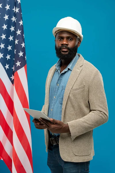 年轻的留胡子的非洲裔美国工程师站在星条旗和蓝色背景的旁边 一边用数字平板电脑 — 图库照片