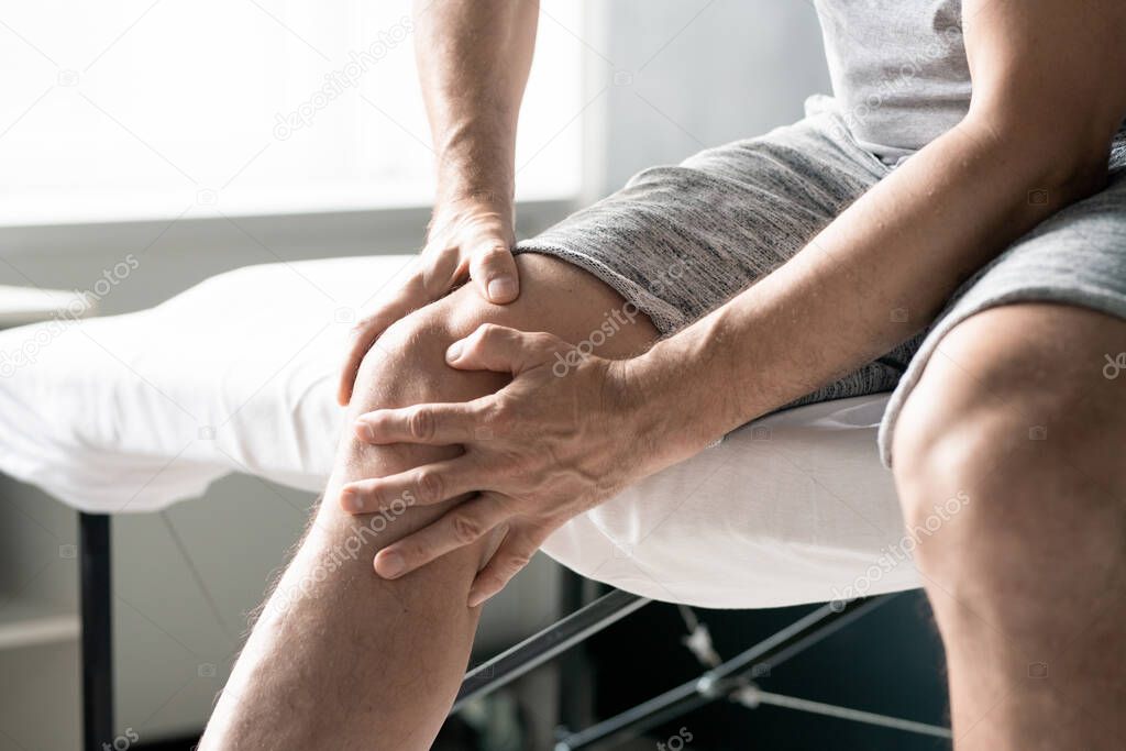 Hands of contemporary mature man massaging his right knee while sitting on table for physiotherapeutic procedures in clinics or medical office