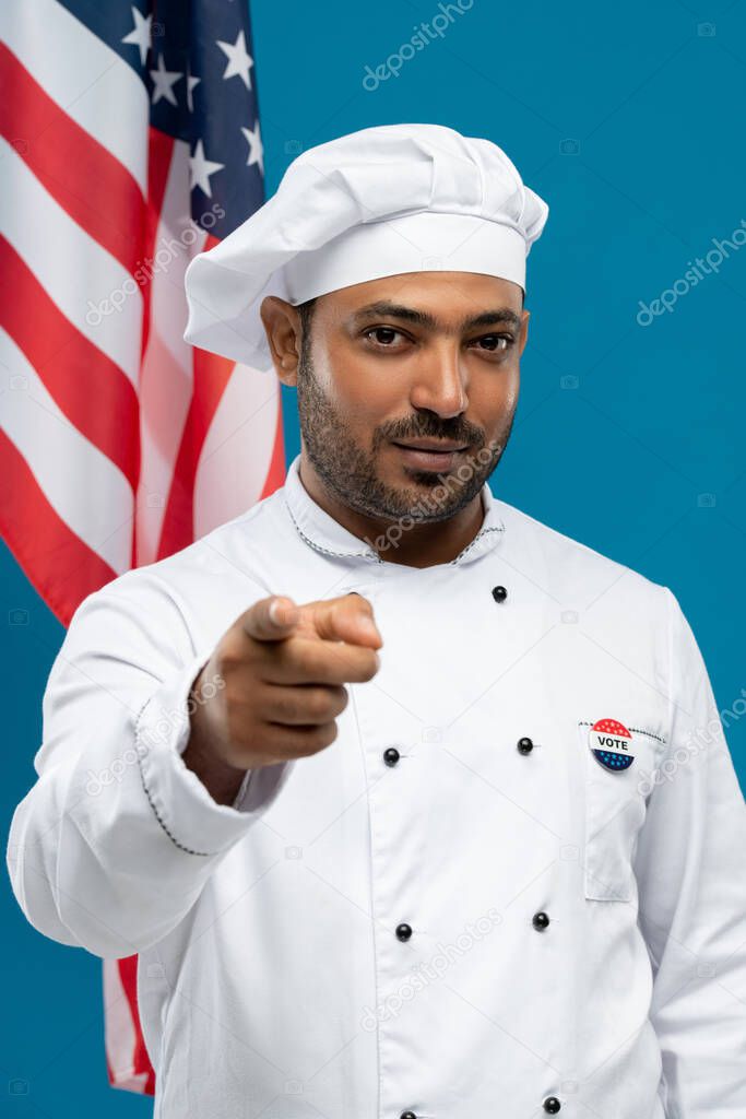 Serious male chef of restaurant in uniform with vote insignia on chest pointing at you while standing against stars-and-stripes flag