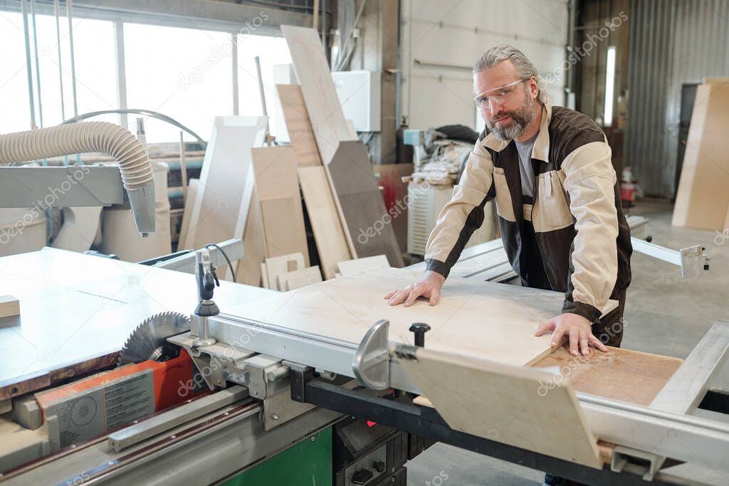 Senior worker of furniture producing factory fixing rectangular board on workbench before cutting or grinding it with electric handtool