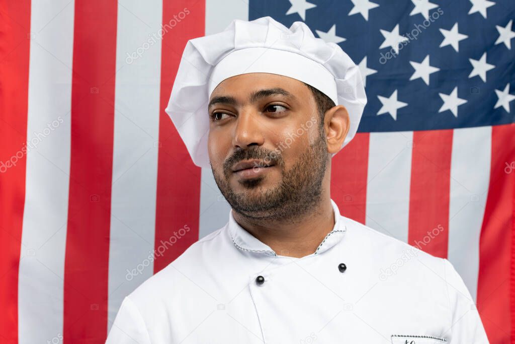 Young professional chef in white cap and coat standing in front of camera against stars-and-stripes background and looking aside