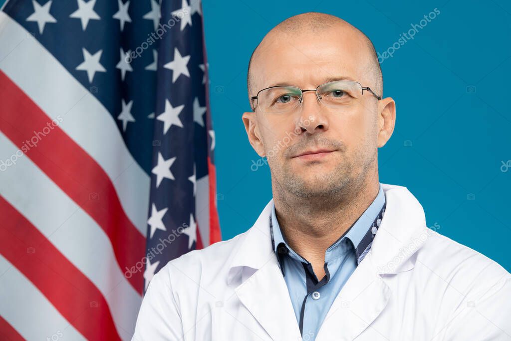 Middle aged bald clinician in eyeglasses and whitecoat looking at you while standing against stars-and-stripes flag in front of camera