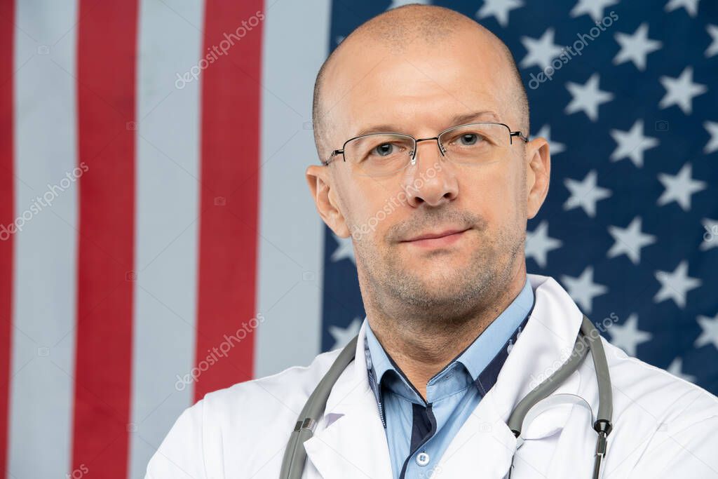 Middle aged contemporary doctor in eyeglasses looking at you while standing against stars-and-stripes flag in front of camera
