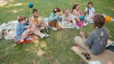 Erkek öğretmenin sakallı, gözlüklü, parktaki battaniyelerde oturan ve dışarıda öğrenirken gezegenlerin modellerini tutan bir grup öğrencinin fotoğrafını çekiyorum.
