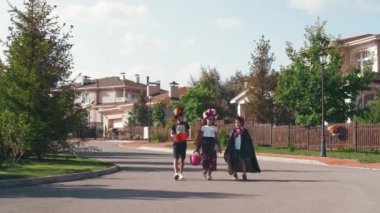 Cadılar bayramı kostümlü, içi şeker dolu sepet ve çanta taşıyan ve banliyöde yürüyen şirin çocukların geniş bir görüntüsü.