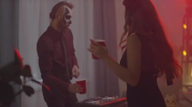 Cadılar Bayramı makyajı yapan, mutlu genç bir kadınla kadeh kaldıran ve DJ 'lik yaparken plastik bardaktan alkol içen bir adamın el kamerası görüntüsü.