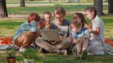 Orta halli mutlu sakallı erkek öğretmenin parkta yeşil çimlerin üzerinde bağdaş kurup dizüstü bilgisayarında meraklı öğrencilere bir şeyler göstermesi.