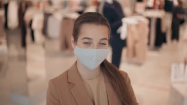 Elbise mağazasında dikilip kameraya bakan maskeli genç bir kadının portresi.