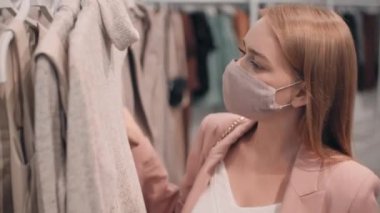 Yüzünde maske olan genç bir kadının elbise rafına doğru yürüdüğü, süvetere baktığı, sonra da onu alıp uzaklaştığı bir fotoğraf.