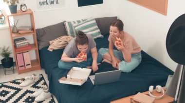 Yüksek açılı mutlu genç kadınların yatak odasında bacak bacak üstüne atarak oturup pizza yerken ve dizüstü bilgisayardan komik bir şeyler izlerken gülüşleri.