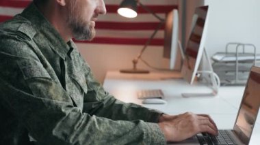 Orta yaşlı, askeri üniformalı bir Amerikan subayının masa başında dizüstü bilgisayar başında otururken çekilmiş yavaş bir görüntüsü.