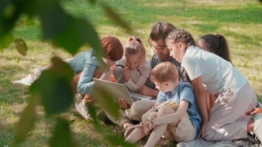 Meraklı okul çocuklarının ve erkek sakallı öğretmenlerinin güneşli yaz gününde parkta öğrenirken dizüstü bilgisayara baktıkları yavaş çekim.