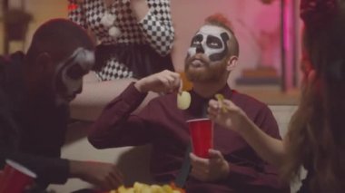 Cadılar Bayramı kostümü giymiş genç erkek ve kadınların ev partisinde kanepede gevşeyip cips yedikleri el kamerası görüntüleri.