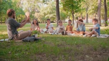 Orta boy sakallı erkek biyolojisi ya da resim öğretmeni parkta yeşil çimlere oturmuş açık hava dersi sırasında küçük öğrencilerinin çizimlerine bakıyor.