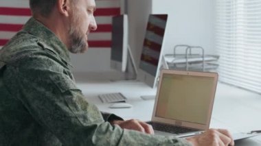 Askeri üniformalı erkek ordu subayının yavaş yavaş takip edilişi. Masada oturup, belgelerle uğraşırken dizüstü bilgisayarda çalışıyor.