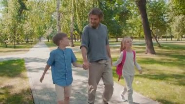 Sakallı genç babanın okul çocuğu ve okul çocuğunun elini tutarken ve güneşli yaz gününde parkta yürürken çekilen fotoğrafı.