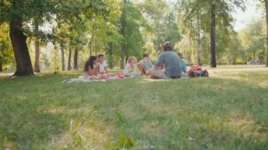 Erkek öğretmenin parkta çimlerin üzerinde battaniyeye oturmuş öğle yemeği yiyen ve onu dikkatle dinleyen bir grup öğrenciyle konuşurken çekilmiş resmi.