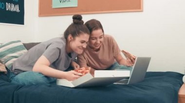 Elde çekilmiş mutlu genç kadınlar yatakta oturup gülerken pizza yerken ve dizüstü bilgisayardan komik bir şey izlerken.