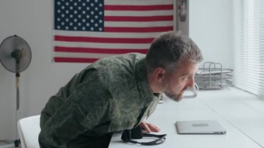 Askeri üniformalı bir Amerikan subayının yavaşça masaya oturup kulaklık takıp dizüstü bilgisayarı açtığı ve çalıştığı bir fotoğraf.