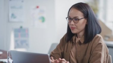 Ofis masasında oturan gözlüklü Asyalı kadının bilgisayarını kullanarak online toplantıya katıldığı orta ölçekli bir fotoğraf.