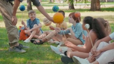 Tanımlanamayan erkek fen öğretmeninin parkta battaniyelerin üzerinde oturan ve güneş sistemini öğrenirken gezegenlerin modellerini tutan sevimli öğrencilerle konuştuğu orta boy bir fotoğraf.