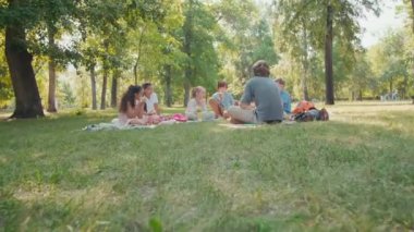 Erkek sakallı öğretmenin battaniyeye oturup öğle yemeği yiyen ve açık hava dersinden sonra dinlenen bir grup mutlu öğrenciyle konuşması.