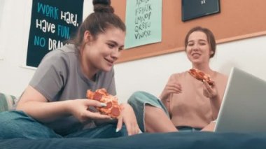 Düşük açılı el kamerasıyla çekilen mutlu genç kadınlar dizüstü bilgisayarda oturup pizza yiyip gülerken ve komik filmler izlerken.