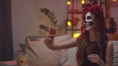 Cadılar Bayramı partisinde koltukta oturup selfie çeken, taç takan ve kafatası makyajı yapan mutlu genç bir kadının yavaş çekimleri.
