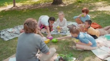 Erkek öğretmenin elinde büyüteçle parkta battaniyeye oturup öğle yemeği yerken görüntüsü.