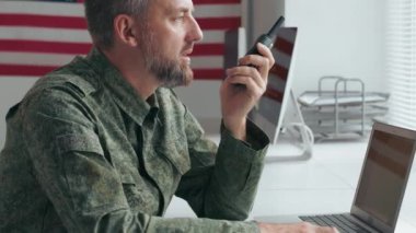 Orta yaşlı, askeri üniformalı Amerikan subayının yavaşça öne eğilip masasında oturup telsizle konuşması.