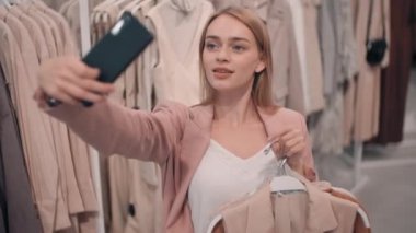 Elbiseleriyle askılık tutan ve mağazada selfie çekerken gülümseyen mutlu genç bir kadının yavaş çekimleri.