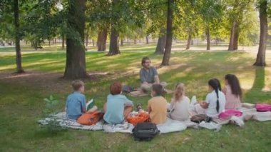 Güneşli bir günde parkta çimlerin üzerinde oturan sakallı ilkokul öğretmeninin el kamerasıyla çekilmiş fotoğrafı ve çocuklara doğa ve çevreyi öğretiyor.