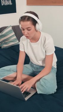 Yurt odasındaki yatakta oturan ve dizüstü bilgisayarda yazarken müzik dinleyen kulaklıklı kızın el kamerasıyla çekilmiş dikey görüntüsü.