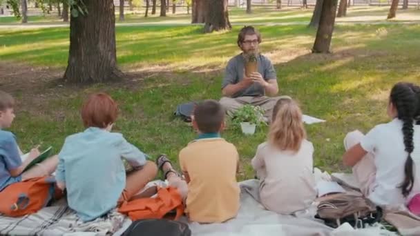 公园里戴着眼镜 双腿交叉坐在毛毯上的大胡子男小学教师和一群好奇的孩子们的植物追踪镜头 — 图库视频影像
