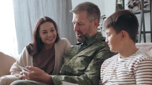 手持慢镜头的快乐的男性退伍军人 留着胡子坐在沙发上 一边分享部署的故事 一边向他快乐的妻子和儿子展示照片 — 图库视频影像