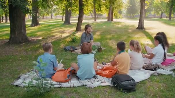 Sakallı Erkek Ilkokul Öğretmeninin Parkta Yeşil Çimlerde Bağdaş Kurup Çocuklara — Stok video