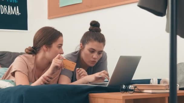 若い女性の追跡ショットは ベッドやオンラインで何かのために買い物に一緒に横たわっています 彼女の友人がノートパソコンを使ってる間に話してる — ストック動画