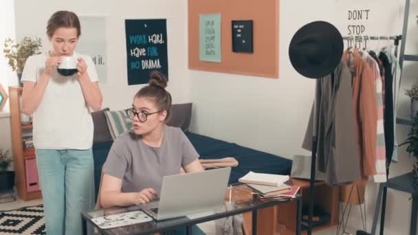 陽気な女の子がお茶を飲んでいるのを追跡し 彼女の女性寮ルームメイトと話して机の上に座ってノートパソコンを入力します 彼女は笑顔で大学の宿題を手伝っています — ストック動画