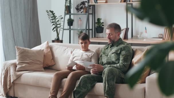 身着军装的男性退伍军人坐在客厅的沙发上 一边给儿子看照片 一边讲述部署情况 — 图库视频影像