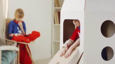 Kız kardeşi telefonda bulanık arka planda oyun oynarken süper kahraman kostümlü genç bir çocuğun beyaz oyuncak karton evden dikizlemesi.