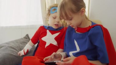 Süper kahraman kostümlü çocukların koltukta oturup heyecanla geçiş seviyesinden bahsettiği orta boy bir fotoğraf.