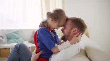 Gülümseyen bir babanın, süper kahraman kostümlü gülen bir kızı kucaklayıp nazikçe dans ettiği ve alınları bükülmüş bir şekilde birbirine baktığı orta boy bir yakın plan.